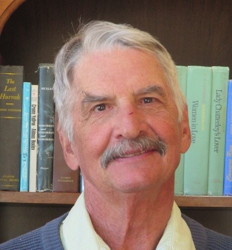 A smiling man with a mustache in front of a bookshelf.