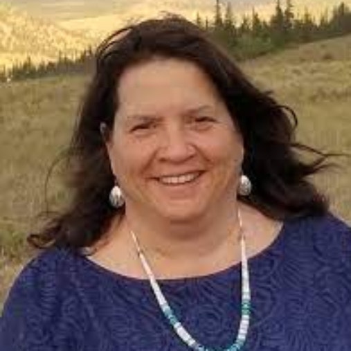 A smiling woman with dark hair, earrings, and a necklace, with a natural landscape in the background.