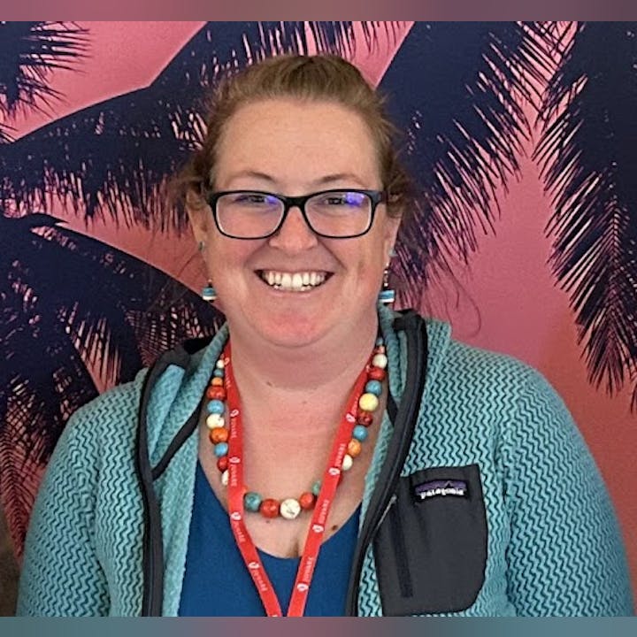 A person wearing glasses and a colorful necklace stands smiling in front of a tropical background with palm silhouettes.
