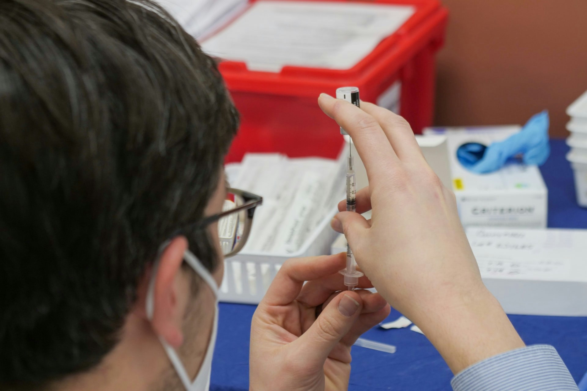 A person preparing a syringe for injection, wearing a mask, with medical supplies in the background.