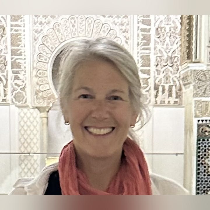 A smiling woman with gray hair and a coral scarf poses against a beautifully detailed background, possibly an architectural site.