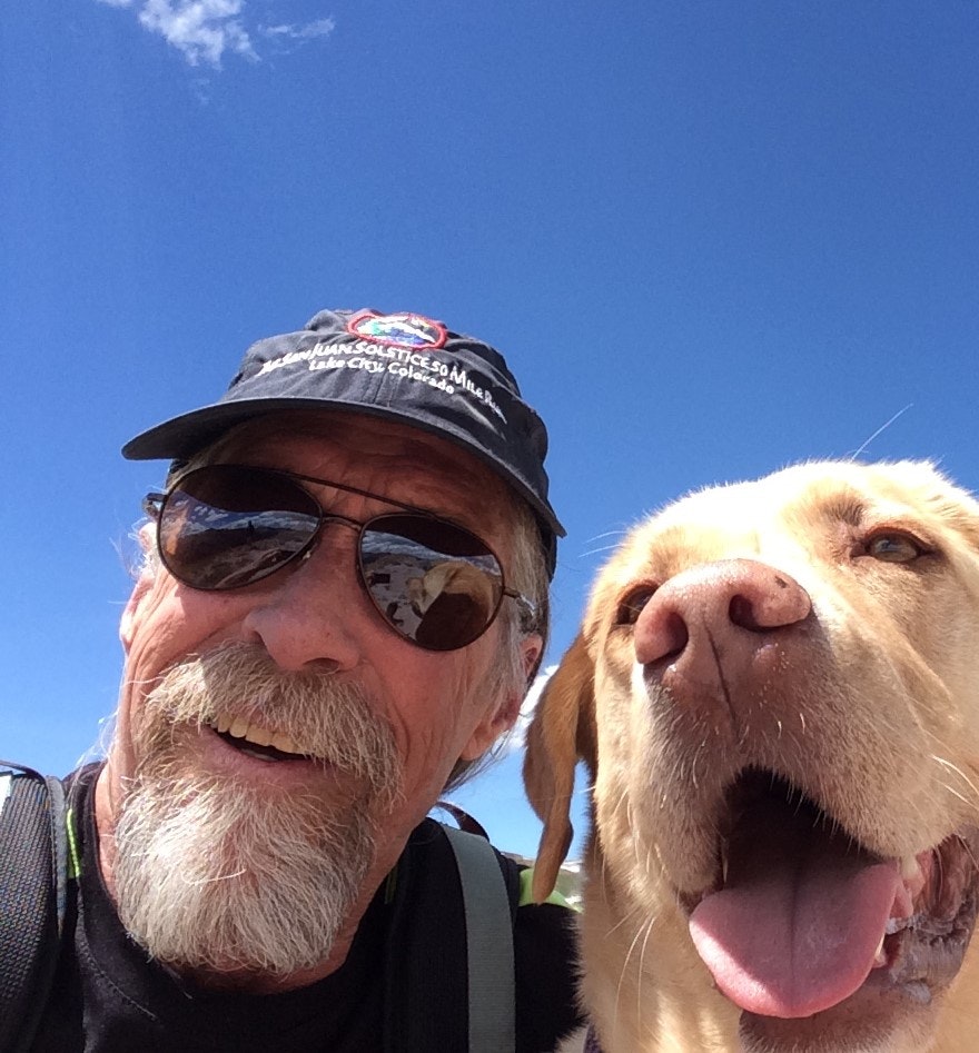 A man with sunglasses and a cap takes a selfie with a happy dog under a clear blue sky.