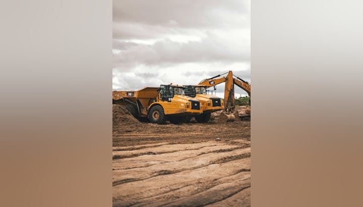 Construction site with two yellow dump trucks and an excavator on dirt ground under cloudy skies.