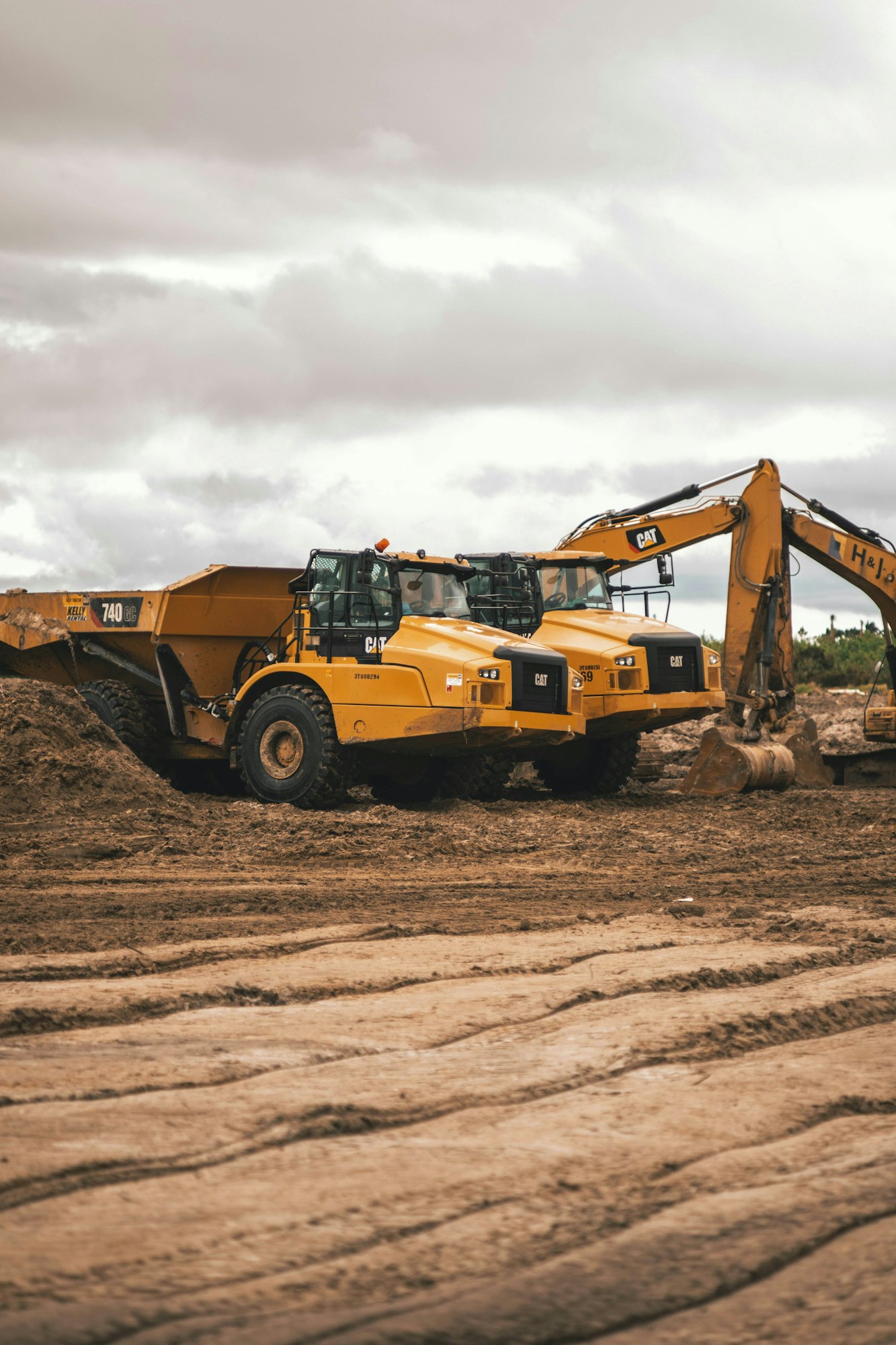 Construction site with two yellow dump trucks and an excavator on dirt ground under cloudy skies.