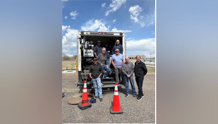 A group of men poses near a truck with equipment, with traffic cones around and a blue sky in the background.