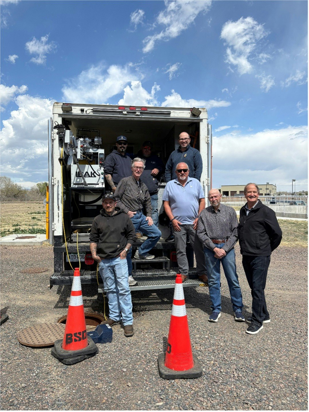 A group of men poses near a truck with equipment, with traffic cones around and a blue sky in the background.