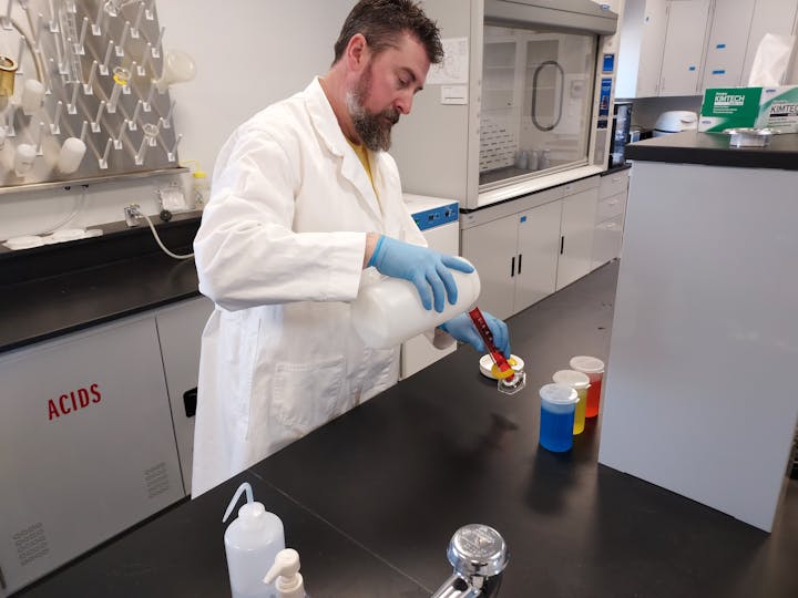 A person in a lab coat and gloves pours liquid from a bottle into a beaker in a laboratory setting.