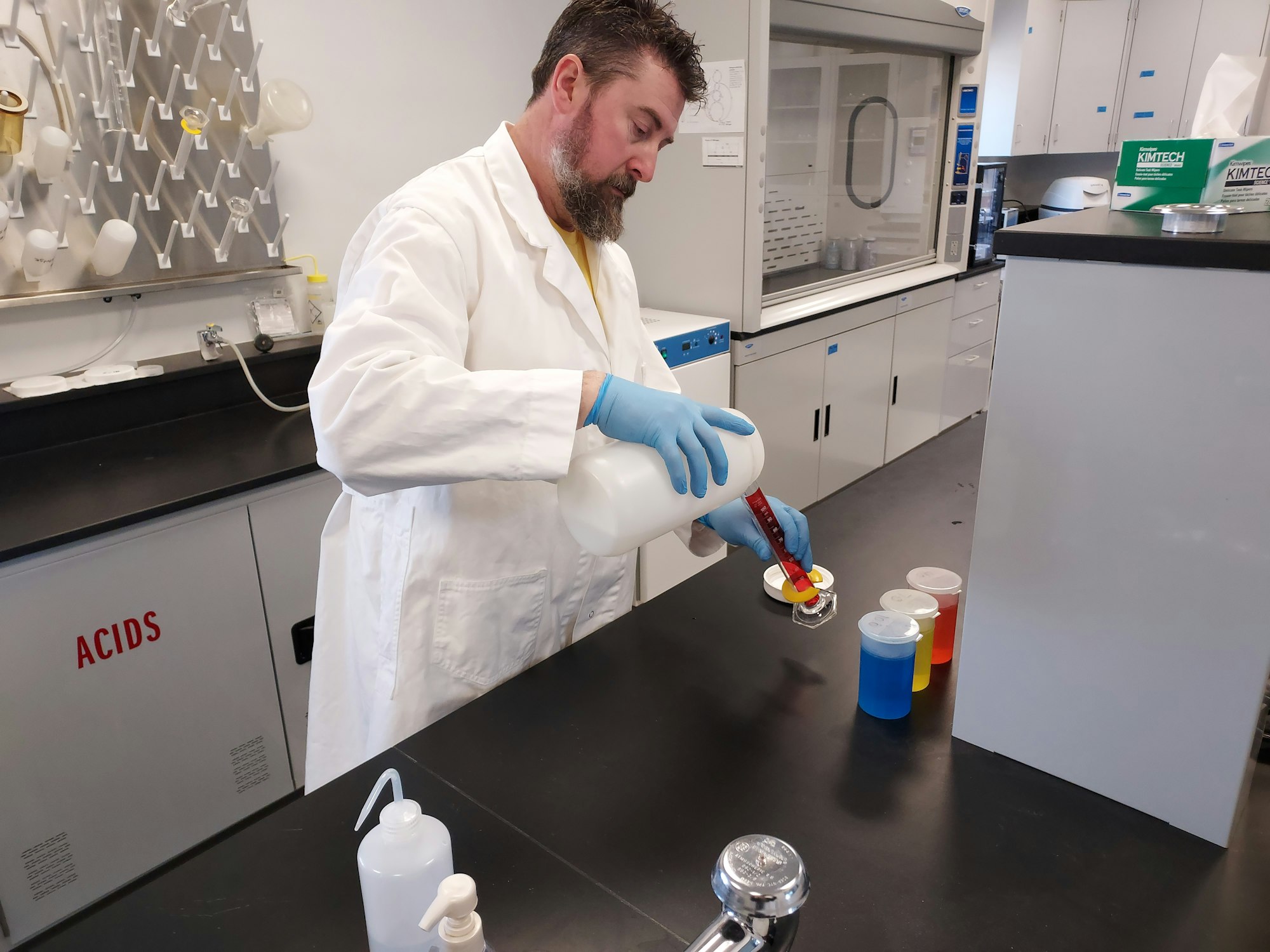 A person in a lab coat and gloves pours liquid from a bottle into a beaker in a laboratory setting.