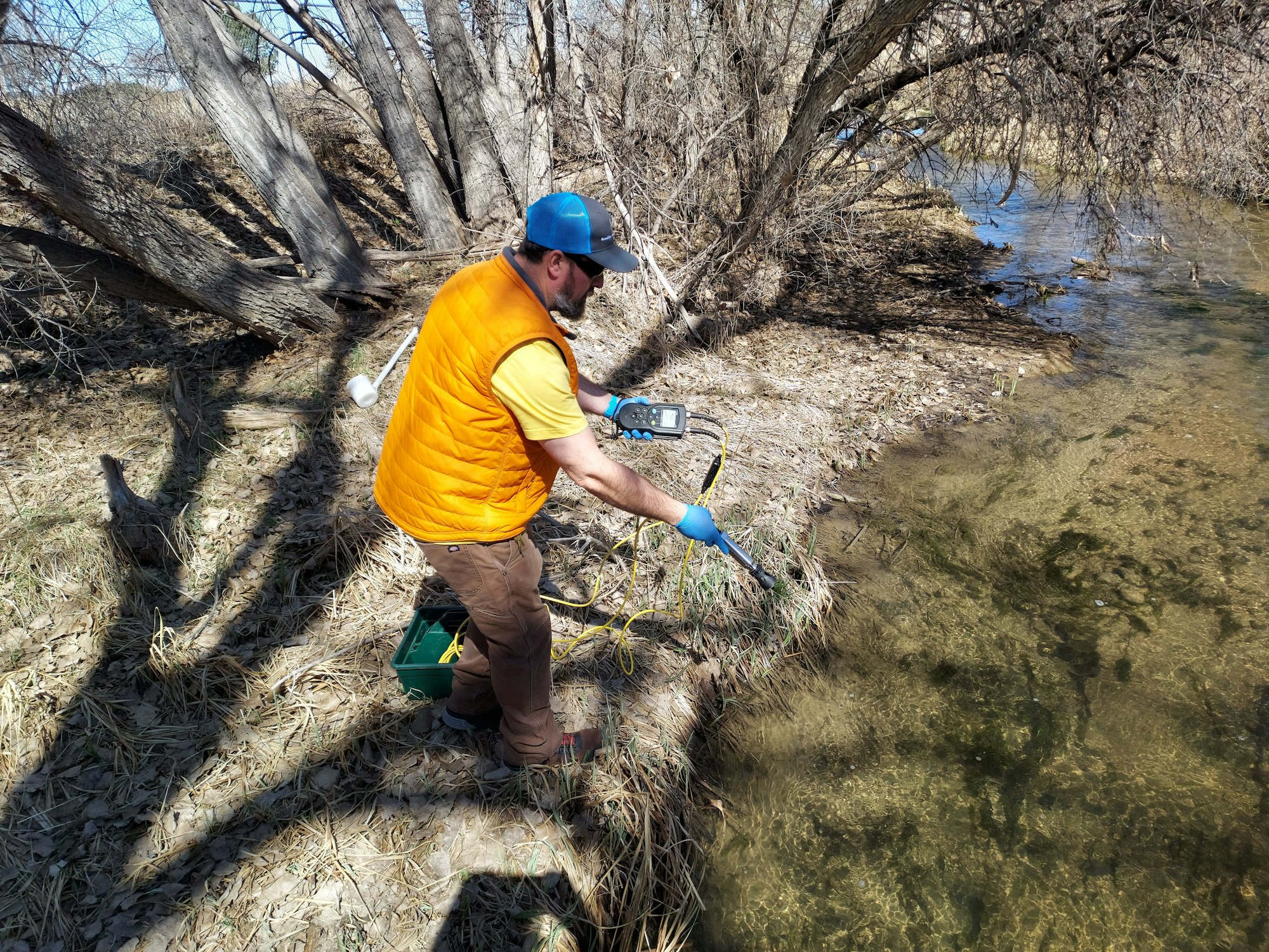 A person in an orange vest with equipment collects water samples at a stream's edge.