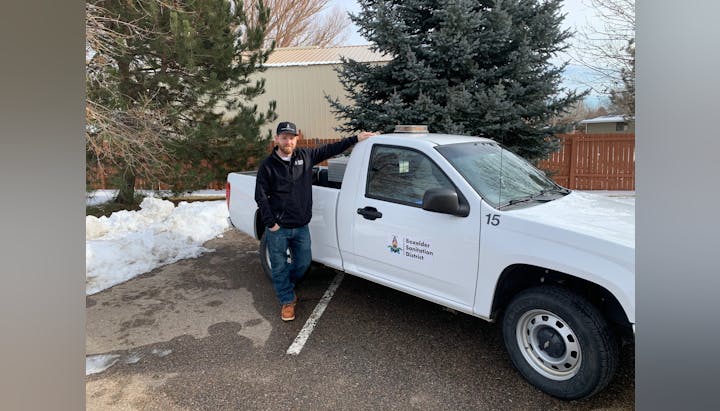 A man standing next to a white truck with the logo "Boxelder Sanitation District" on it, trees and snow are visible.