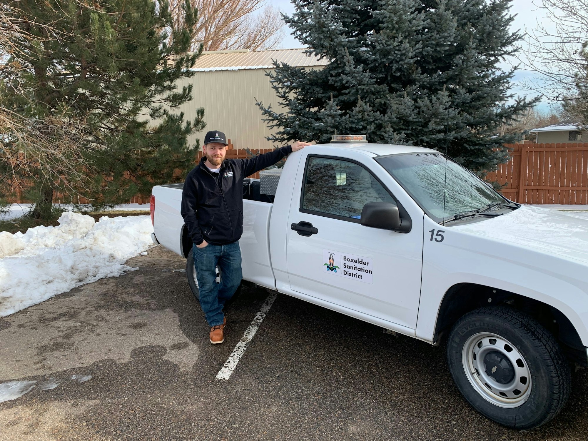 A man standing next to a white truck with the logo "Boxelder Sanitation District" on it, trees and snow are visible.