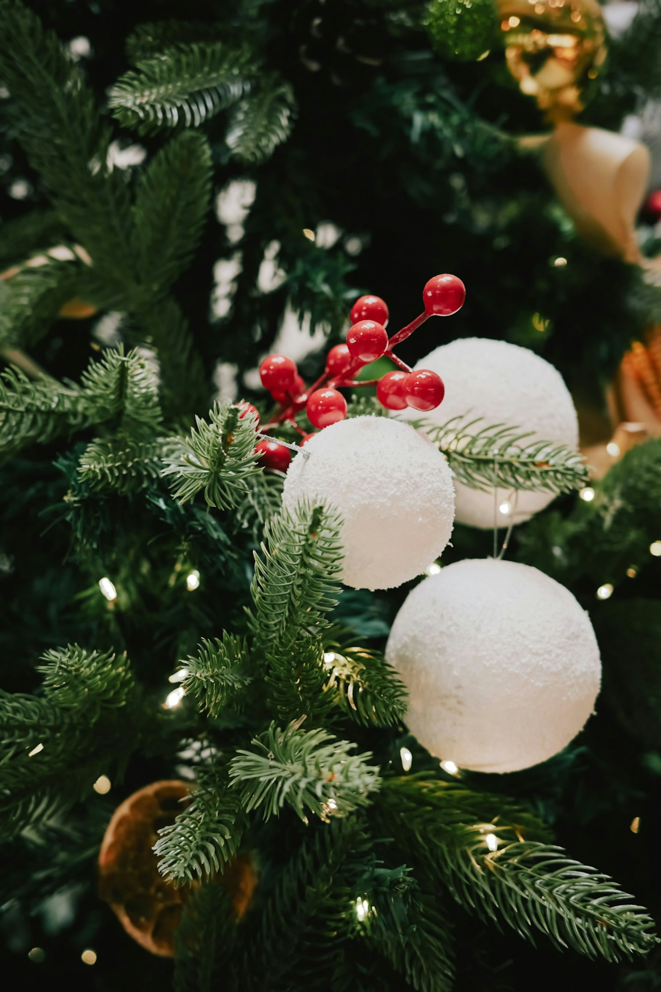 A close-up of a Christmas tree featuring white decorations, red berries, and green foliage, with soft glowing lights.