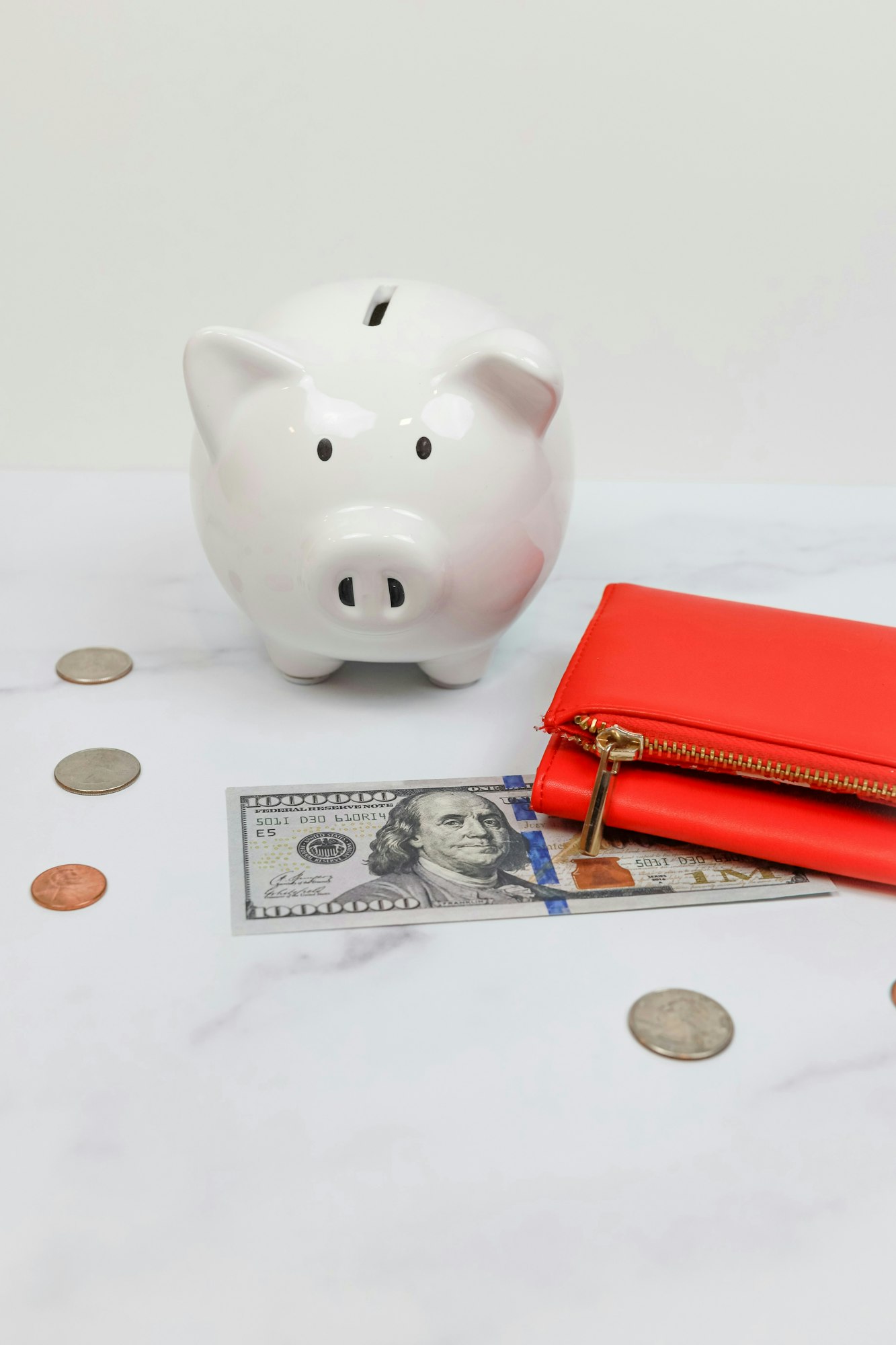 A white piggy bank, a red wallet, a $100 bill, and several coins on a marble surface.