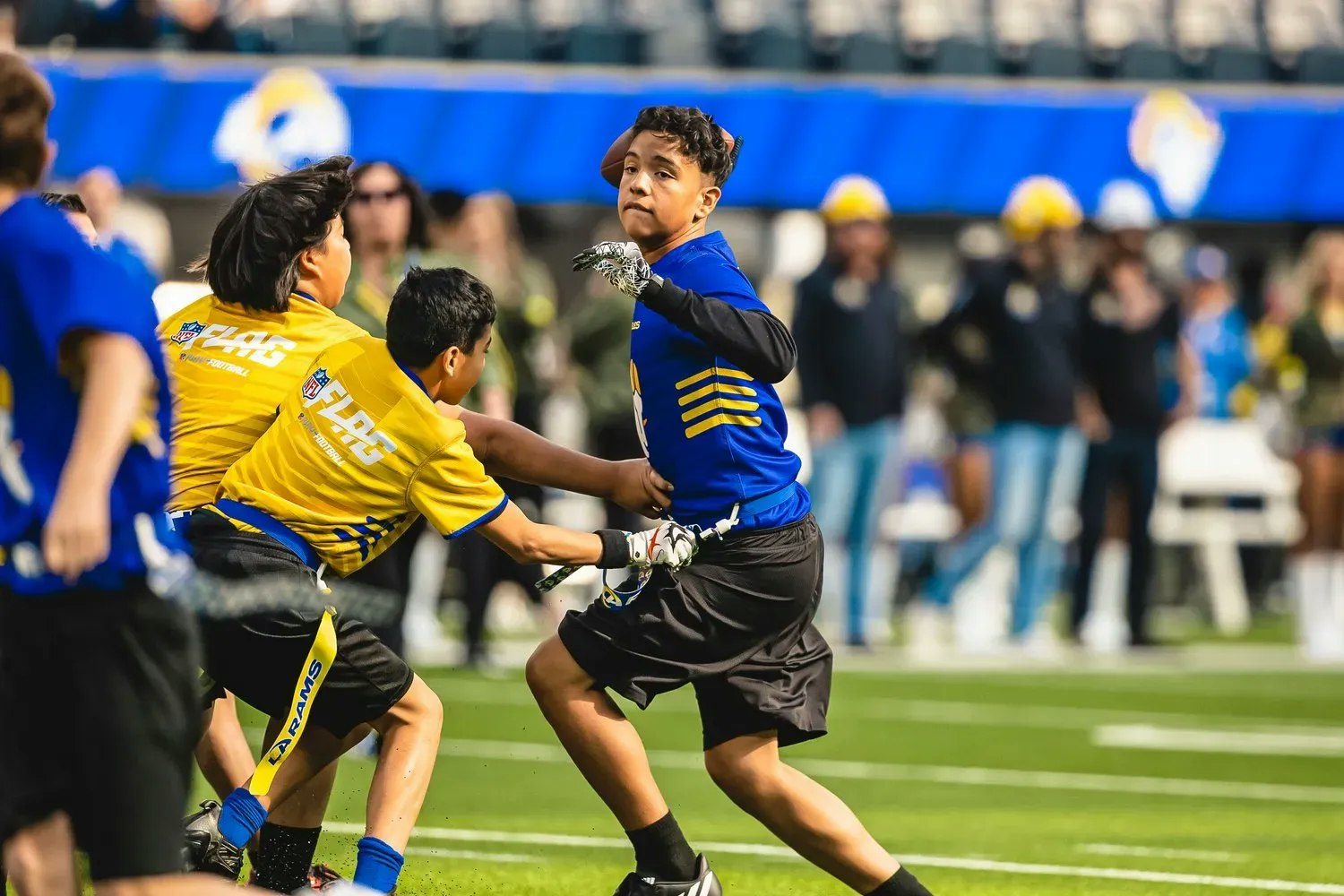 Kids are playing flag football, wearing blue and yellow jerseys, on a grassy field with spectators in the background.