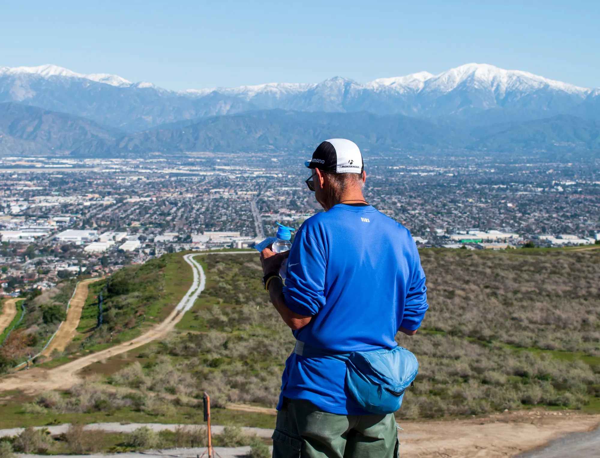 A person in a blue shirt stands on a hill, looking out over a city and snow-capped mountains in the distance.