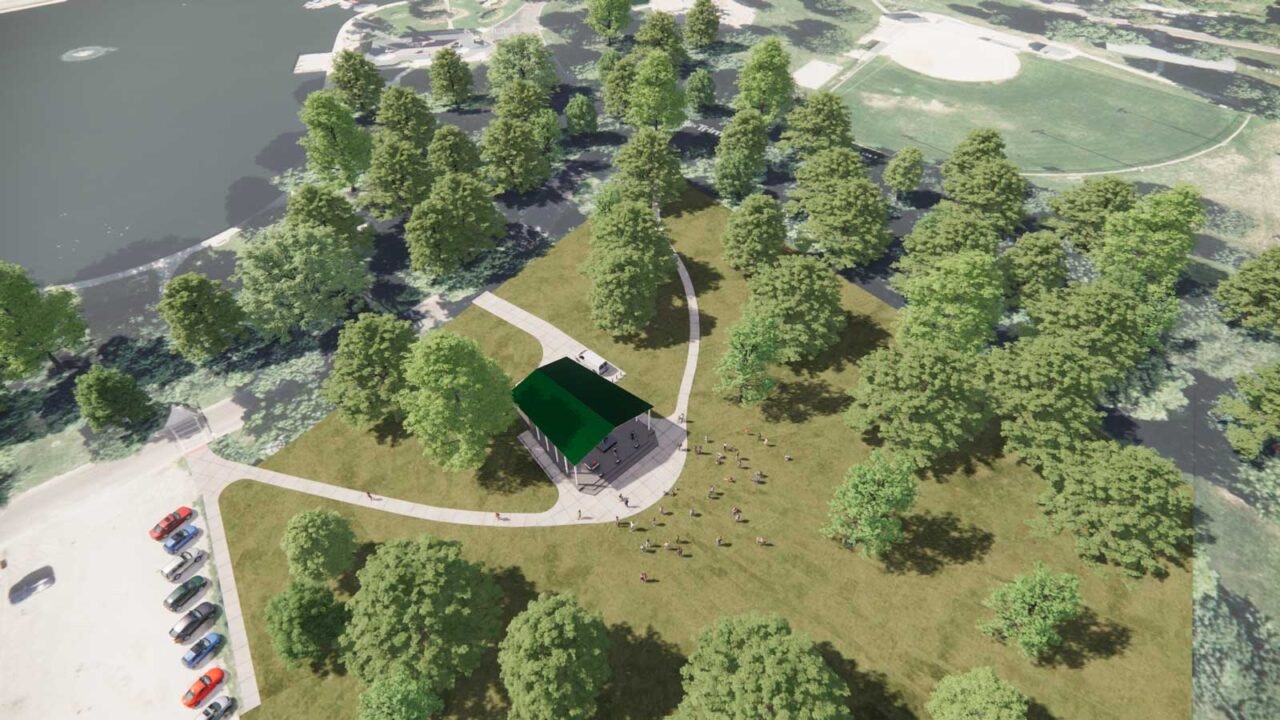 An aerial view of a park with a pavilion surrounded by trees and a parking area, near a water body and sports fields.