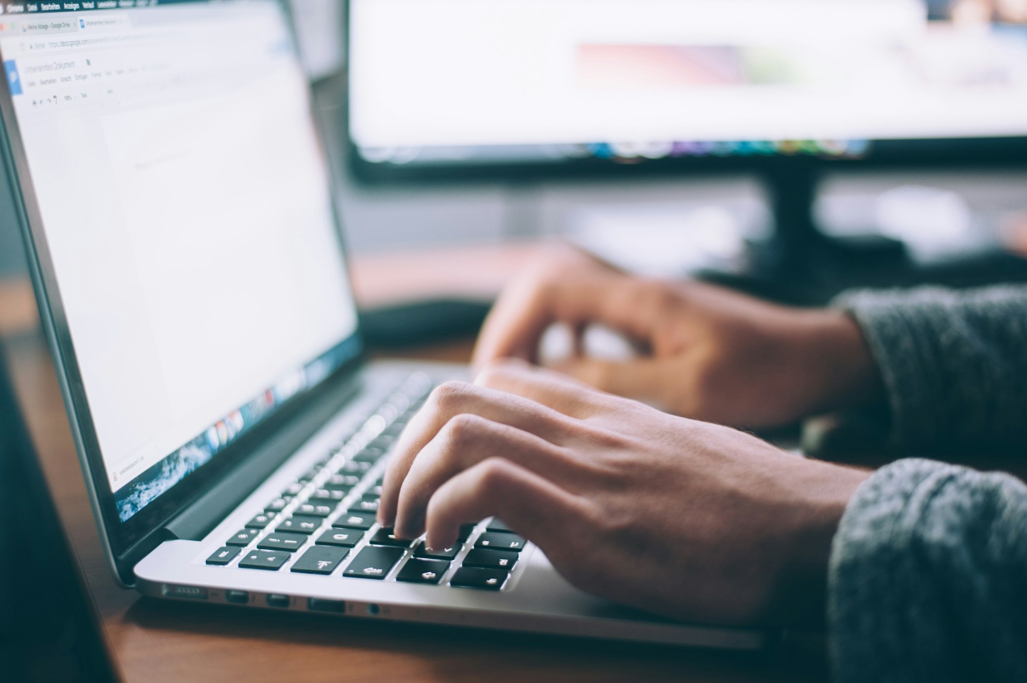 A person typing on a laptop, with another monitor visible in the background, likely working on a document or project.