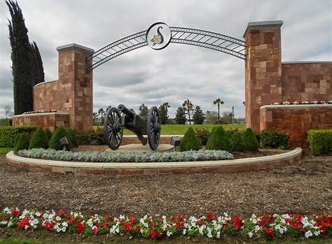 A brick entrance sign with an archway, a cannon, and landscaped flowers in front.
