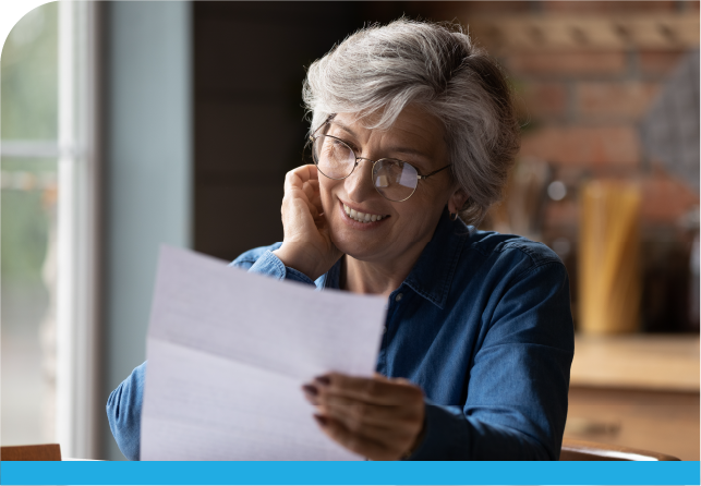 A woman with gray hair and glasses smiles while reading a letter at a table, conveying a sense of joy and engagement.
