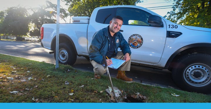 A man kneels beside a truck, taking notes while inspecting a spot in the ground, likely for a maintenance or planting task.