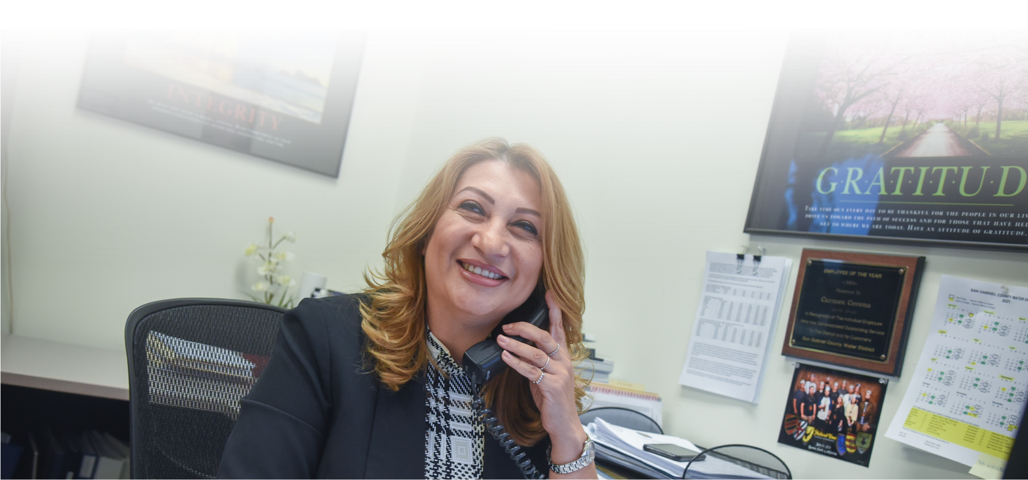A smiling woman at a desk talking on the phone, surrounded by motivational posters and office materials.