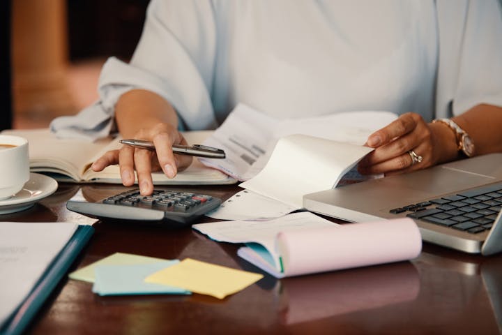 A person manages finances using a calculator, laptop, and papers, with a cup of coffee nearby.