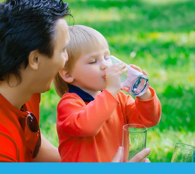 A man and a young child enjoy a sunny day outside, with the child drinking from a glass while the man smiles beside him.