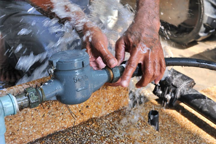 A person is fixing a water meter, with water splashing around their hands and tools, highlighting plumbing work.