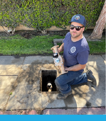 A man kneels by a hole in the ground, holding a water meter while wearing sunglasses and a cap. Green bushes in the background.