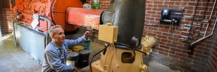 A technician inspects machinery in an industrial setting, surrounded by pipes, gauges, and equipment on a brick wall.