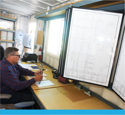 A man is working at a desk with large printed plans displayed on boards in a well-lit workspace.