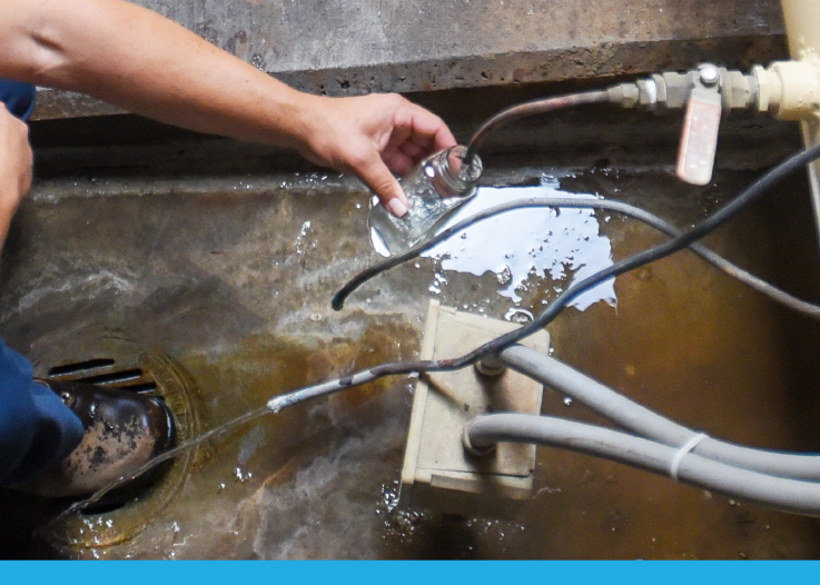 A person is holding a clear bottle over a waterlogged area with pipes and a sump pump visible in the environment.