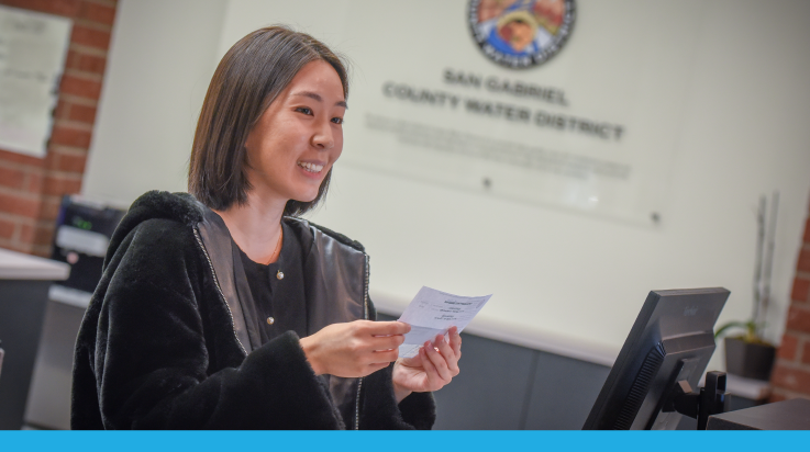 A woman is smiling and holding a paper while standing at a reception desk with a "San Gabriel County Water District" sign in the background.