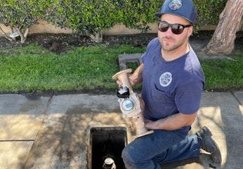 A person in a cap and sunglasses holds a water meter above an open pit, with grass and trees in the background.