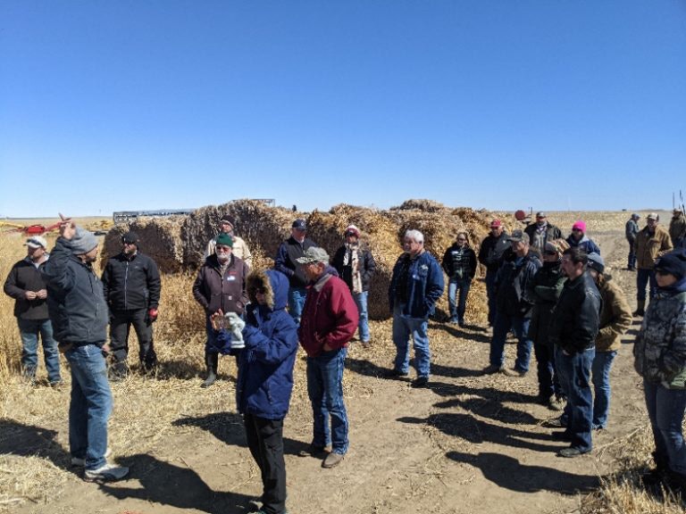 A group of people is gathered outdoors, likely for a presentation or discussion, with straw bales in the background.
