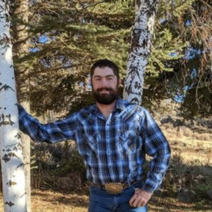 A man in a plaid shirt and jeans standing by birch trees.