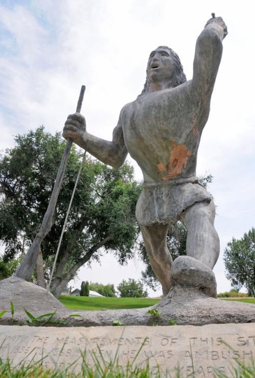 A statue depicting a historical figure holding a spear, with an arm raised, set against a background of trees and grass.