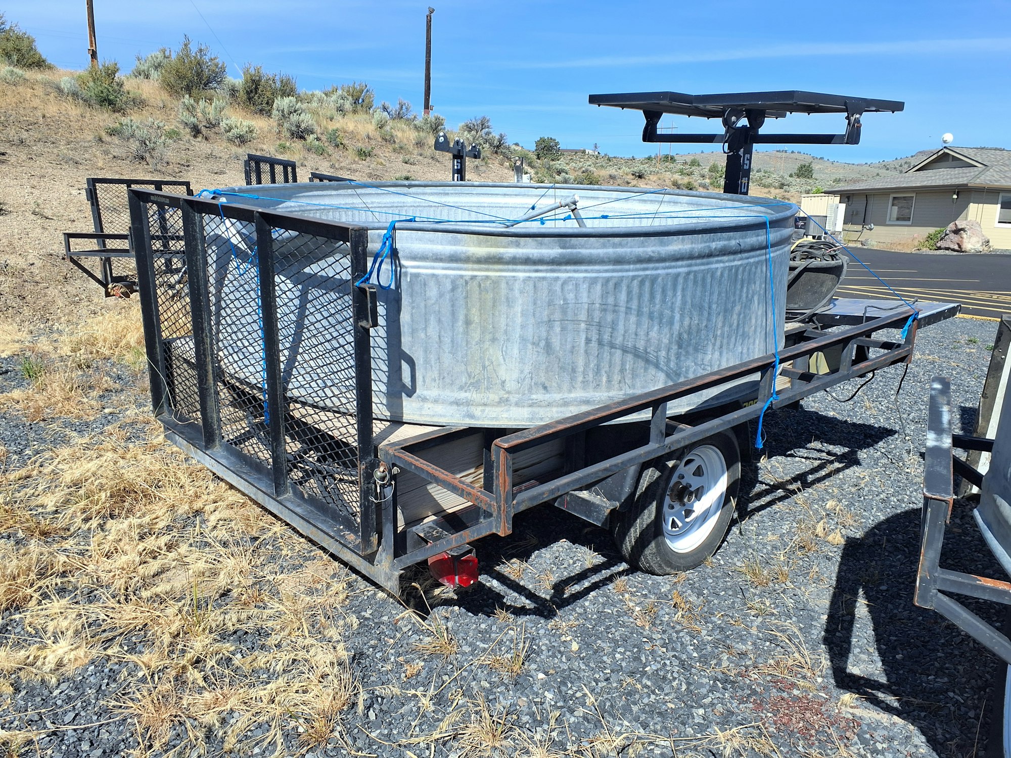 A metal water tank on a trailer, secured with blue straps, parked on rocky ground with some dry grass and a distant building.