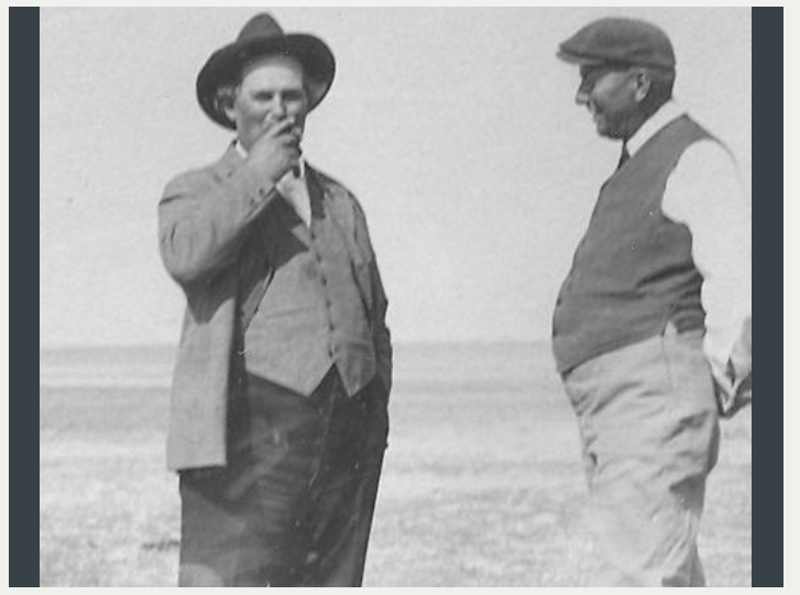 Two men standing outdoors, one smoking, both in early 20th-century attire.