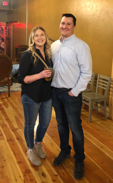A smiling man and woman standing together indoors, with the woman holding a drink.