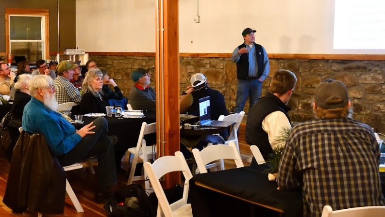 A group of people sits at tables, listening to a speaker presenting in a warmly lit venue.