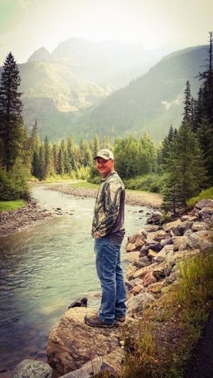 A person standing by a mountain river, surrounded by trees and mountains in the background.