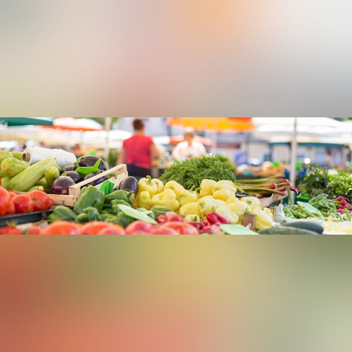 A variety of fresh vegetables displayed at a market stall with blurred shoppers in the background.