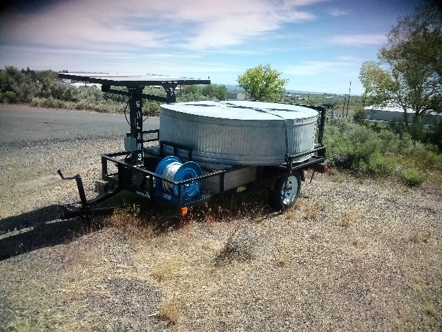 A water tank on a trailer with a blue hose reel, parked in a dry grassy area under a clear sky.