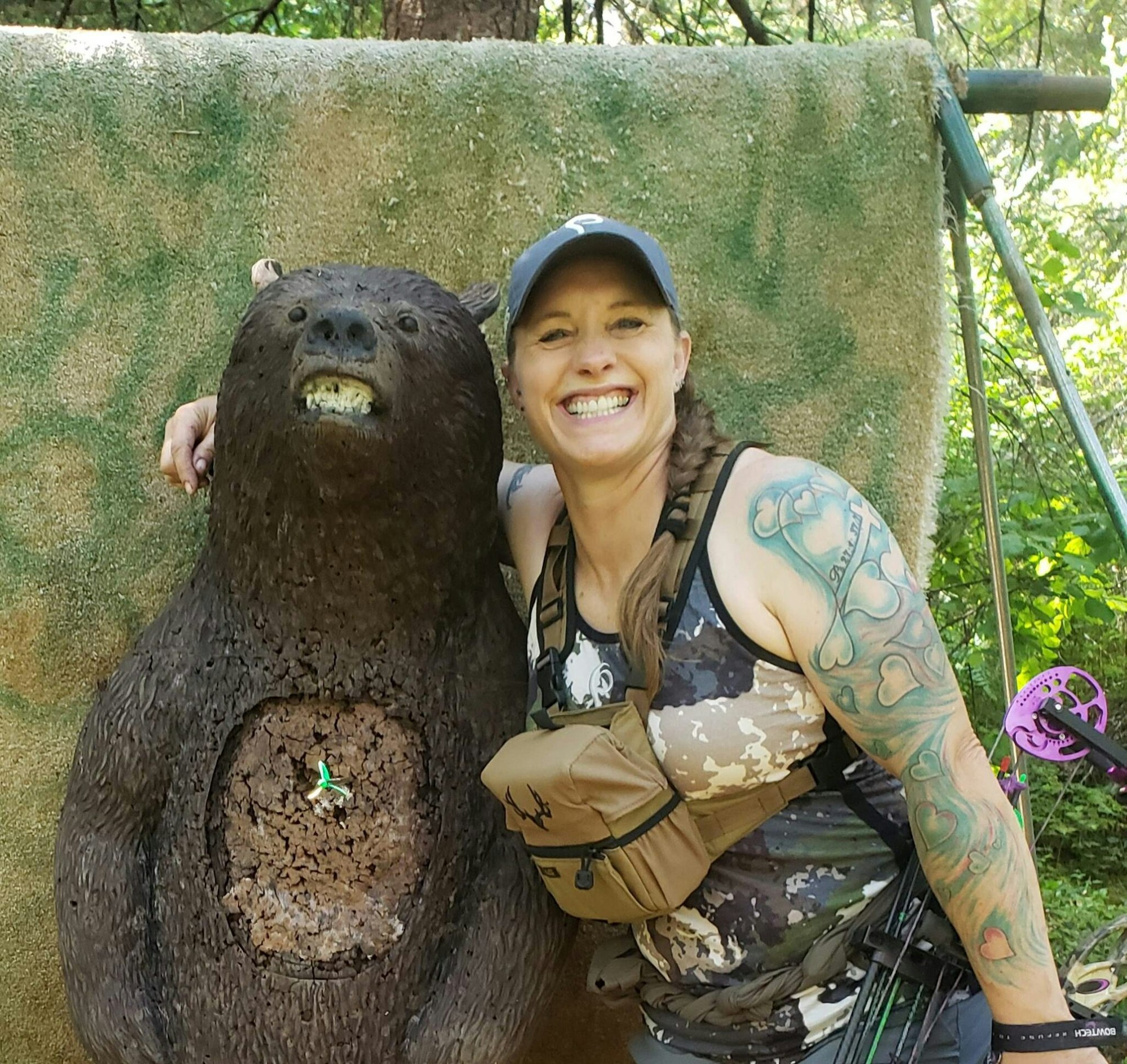 A smiling woman with tattoos posing next to a bear archery target with an arrow hit.