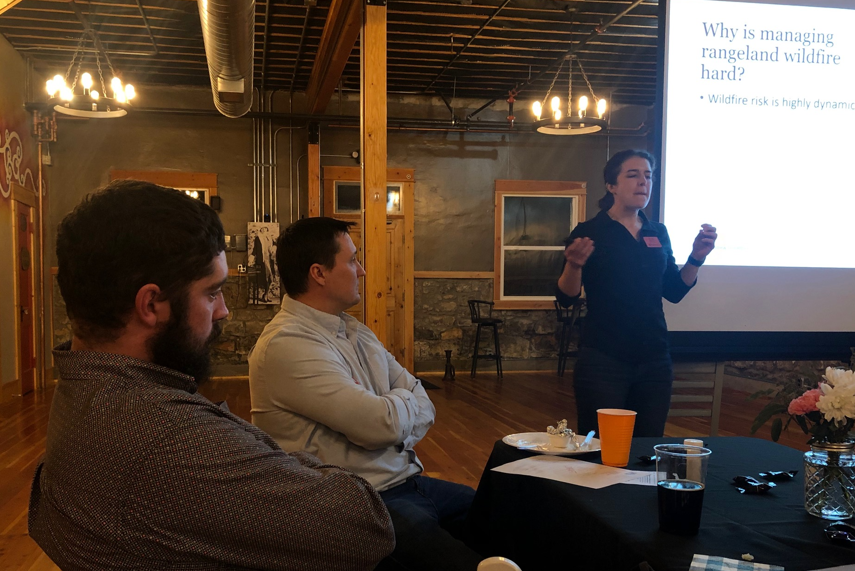 A presentation in progress with attendees listening and a speaker discussing rangeland wildfire management.