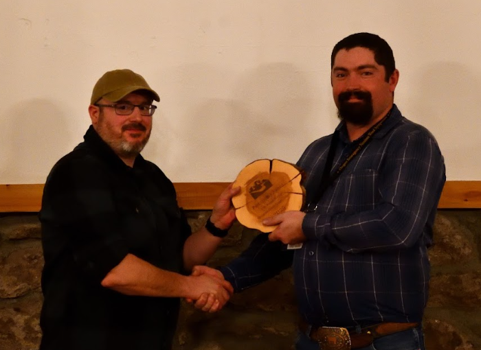 Two men are shaking hands; one is handing a wooden plaque to the other in a casual setting.