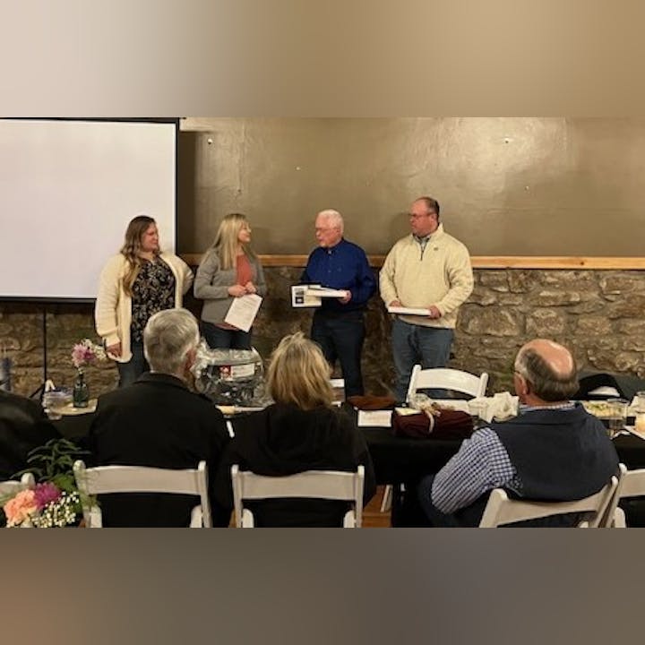 Four people standing, one receiving an award/document, in a room with seated audience.