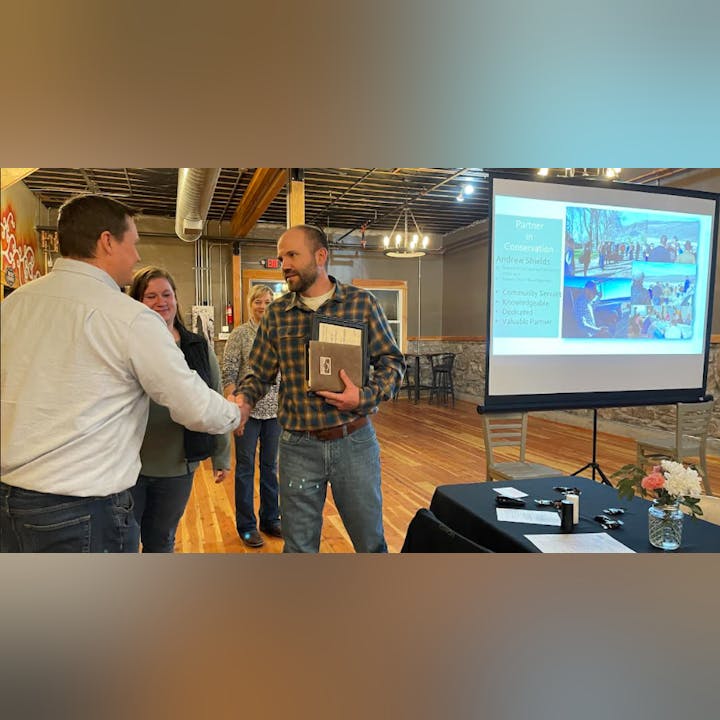 Two people shaking hands at an event, a presentation screen in the background highlights a conservation partnership.
