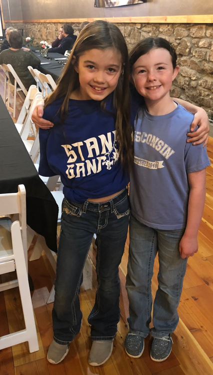 Two smiling children standing together indoors, one wearing a "STRANGER" themed blue T-shirt and the other in a gray "WISCONSIN" T-shirt.